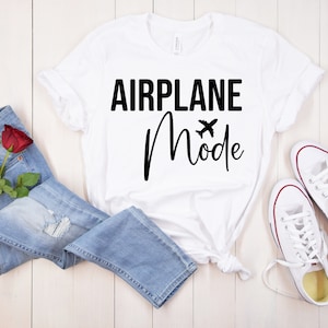 May include: White t-shirt with black text that reads "Airplane Mode" with a small airplane icon. The shirt is laid out on a wooden surface with a pair of white sneakers and blue jeans.