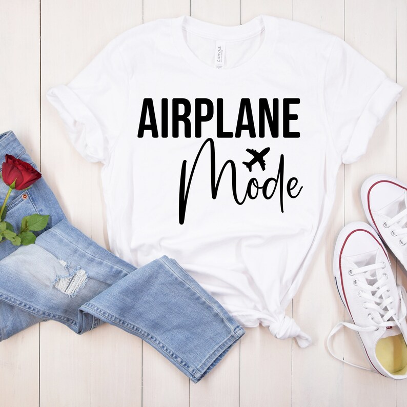 May include: White t-shirt with black text that reads "Airplane Mode" with a small airplane icon. The shirt is laid out on a wooden surface with a pair of white sneakers and blue jeans.