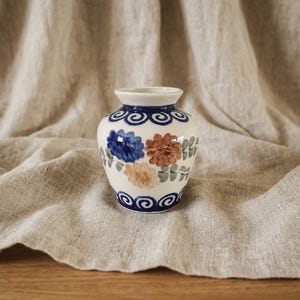 Pode incluir: Um pequeno vaso de cerâmica com base na cor creme, apresentando motivos florais pintados à mão em azul e castanho, folhas verdes e padrões de espirais azul-escuro ao redor do pescoço e da base, com acabamento liso e brilhante e corpo arredondado.