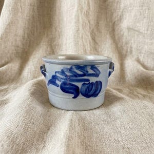 Pode incluir: Um pote de cerâmica cinza-claro com duas pequenas alças laterais e um design abstrato de pinceladas em azul cobalto pintado à mão que envolve o exterior. O recipiente tem uma abertura circular larga e um acabamento liso e fosco.