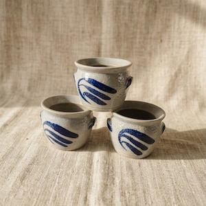Può includere: Tre piccoli vasi in ceramica color bianco sporco maculato, ciascuno dotato di due manici laterali. Ogni vaso presenta un motivo astratto a forma di foglia o piuma blu scuro dipinto a mano sulla parte anteriore. Hanno una forma arrotondata.