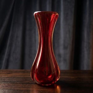May include: A tall, translucent ruby red glass vase with a smooth, curved silhouette. The vase features a narrow neck that flares outward at the top and a rounded, bulbous base with vertical indentations that create a ribbed, petal-like effect.