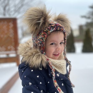 May include: A colourful knitted winter hat with two large faux fur pom-poms. The hat has ear flaps and braided ties. The child is wearing a navy blue jacket with white heart-shaped designs and a cream-coloured scarf.