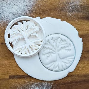May include: A white plastic cookie cutter and a white imprint of a tree with roots in a white dough. The cookie cutter is shaped like a tree with roots.