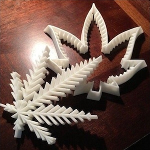 Könnte beinhalten: Zwei weiße, 3D-gedruckte Marihuana-Blattdesigns. Ein Design ist eine traditionelle Marihuana-Blattform, das andere ist ein detaillierteres Design, das an einen Farn erinnert.