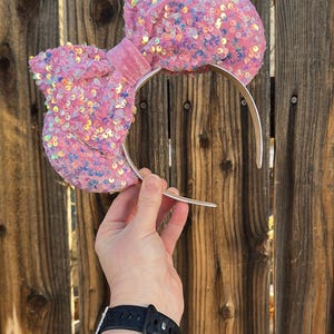 May include: A pink sequined bow headband with a silver band. The bow is large and fluffy, covered in iridescent sequins. The headband is held up against a wooden fence.