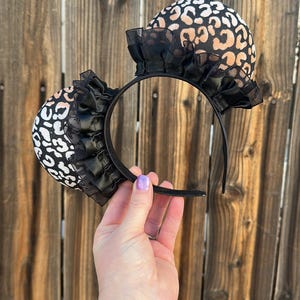 May include: Black headband with two ears featuring a leopard print design. The ears are adorned with black ruffles. The headband is held up against a wooden fence.