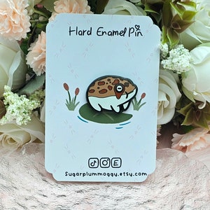 Peut inclure: Une épingle en émail dur représentant une grenouille de dessin animé avec des marques marron et blanches, assise sur un nénuphar. L'épingle est sur une carte blanche avec le texte "Hard Enamel Pin" et des icônes de médias sociaux. Le fond comprend des fleurs.