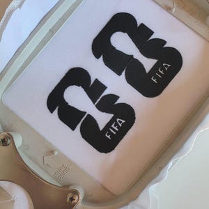 May include: White fabric with the black embroidered number "23" and the word "FIFA". The fabric is held in a metal embroidery hoop. The design is centered on the fabric.