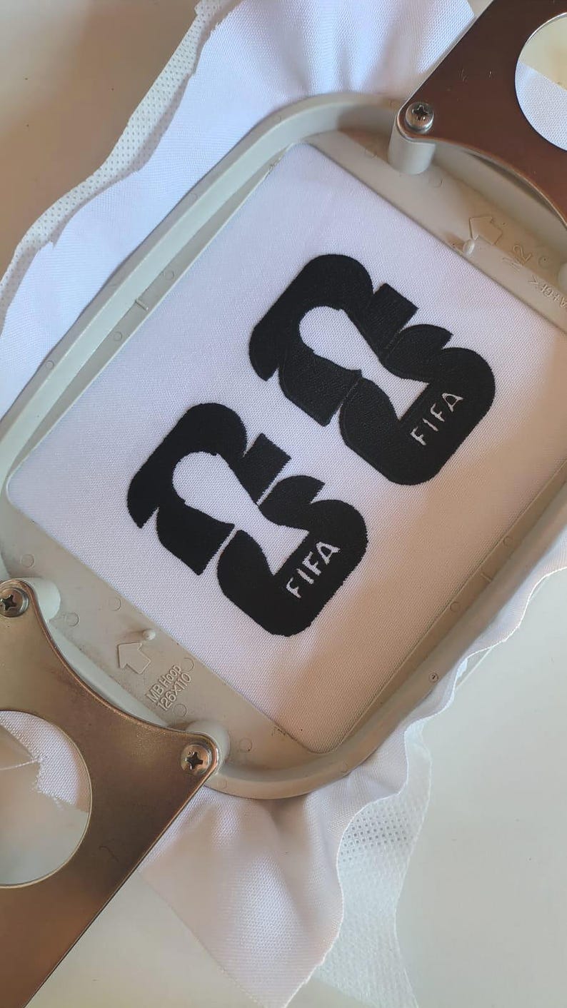 May include: White fabric with the black embroidered number "23" and the word "FIFA". The fabric is held in a metal embroidery hoop. The design is centered on the fabric.