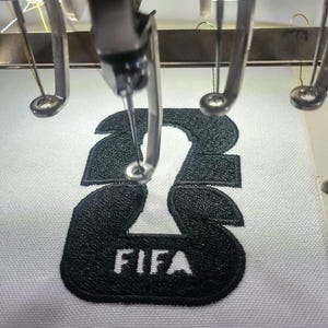 May include: Close-up of an embroidery machine stitching a black number "23" with the word "FIFA" below it onto white fabric. The machine's needle is positioned over the design, creating a detailed, textured finish.