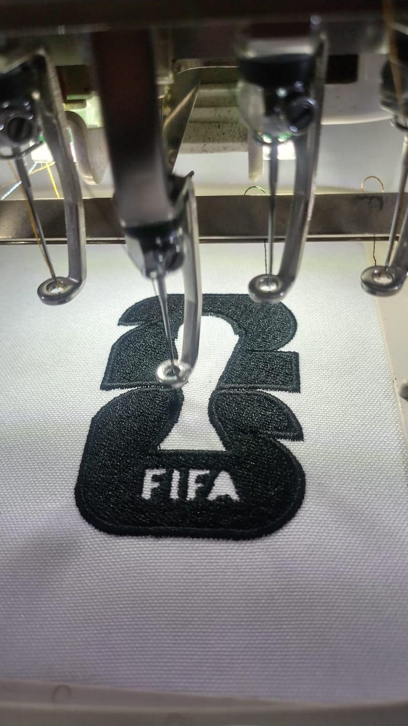 May include: Close-up of an embroidery machine stitching a black number "23" with the word "FIFA" below it onto white fabric. The machine's needle is positioned over the design, creating a detailed, textured finish.