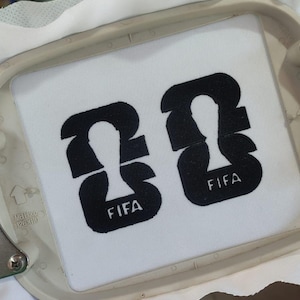 May include: Two black embroidered FIFA logos on a white fabric, set within an embroidery hoop. The logos feature a stylized figure and the word "FIFA" in a clean, sans-serif font. The embroidery machine is visible in the background.