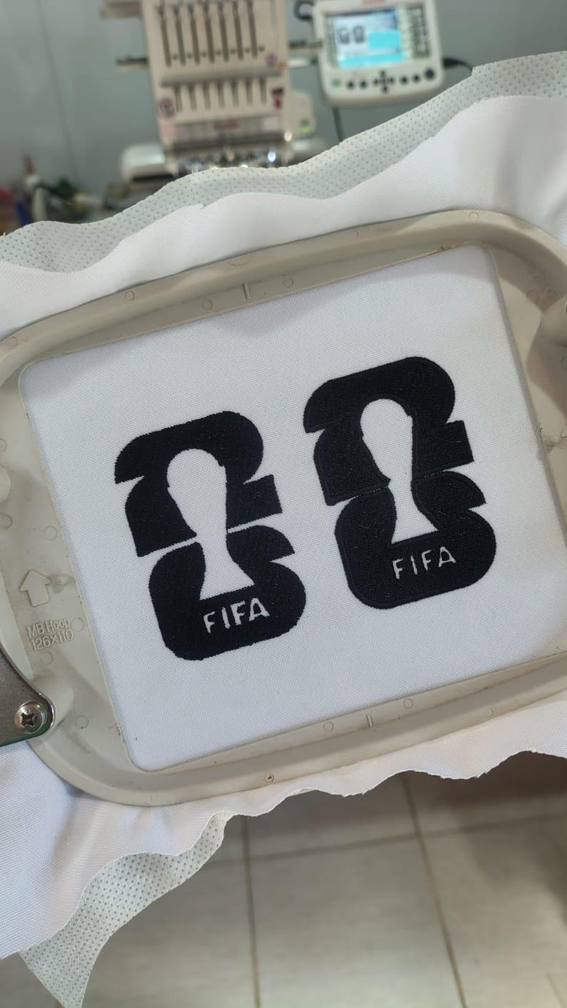 May include: Two black embroidered FIFA logos on a white fabric, set within an embroidery hoop. The logos feature a stylized figure and the word "FIFA" in a clean, sans-serif font. The embroidery machine is visible in the background.