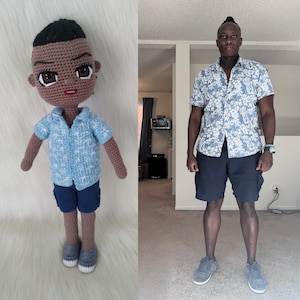 May include: A crocheted doll wearing a blue and white patterned shirt and blue shorts. The doll has brown skin and black hair. The doll is standing on a white fluffy surface. In the background, a person is wearing a blue and white patterned shirt and blue shorts. The person is standing on a light brown floor.