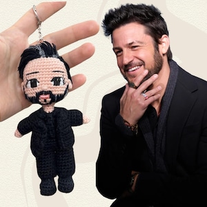May include: A handmade crochet doll keychain featuring a man with black hair and a beard, wearing a black suit. The doll is held up by a silver keychain. The background is a light beige color.