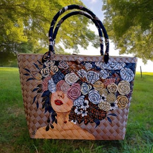 Può includere: Borsa tote intrecciata con un design floreale raffigurante il volto di una donna. La borsa ha un corpo intrecciato marrone, manici neri e blu e un'illustrazione colorata di fiori e del volto di una donna. La borsa è su uno sfondo verde.