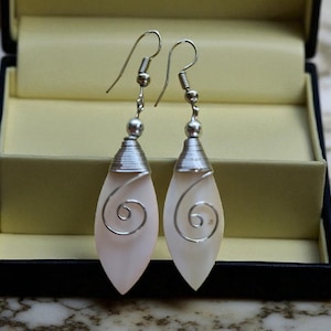 May include: Dangle earrings with a pale pink, teardrop-shaped design. The earrings have silver-coloured hooks and a silver wire spiral. They are presented in a black jewellery box with a light yellow interior.