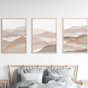 May include: Three framed prints with abstract watercolor paintings of brown and beige mountains. The prints are hung on a white wall above a bed with gray bedding.