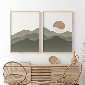 Op de afbeelding: Twee ingelijste abstracte landschapsdrukken met groene en bruine bergen en een zon in de rechter print. De prints hangen aan een witte muur boven een houten kast met twee rieten stoelen en een rieten kruk.
