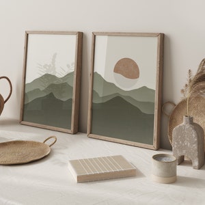Op de afbeelding: Twee ingelijste abstracte landschapsdrukken in een groen en bruin kleurenschema. De prints hangen aan een witte muur en tonen een gestileerd berggebied en een zon.