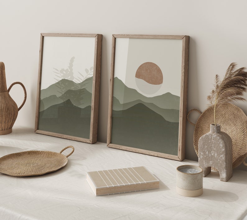 Op de afbeelding: Twee ingelijste abstracte landschapsdrukken in een groen en bruin kleurenschema. De prints hangen aan een witte muur en tonen een gestileerd berggebied en een zon.