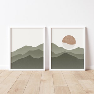 Op de afbeelding: Twee ingelijste prints met een minimalistisch ontwerp met een landschap van groene bergen en een enkele oranje zon aan de hemel.