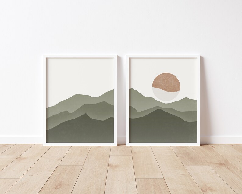 Op de afbeelding: Twee ingelijste prints met een minimalistisch ontwerp met een landschap van groene bergen en een enkele oranje zon aan de hemel.