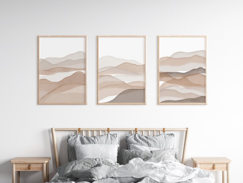May include: Three framed prints with abstract watercolor paintings of brown and beige mountains. The prints are hung on a white wall above a bed with gray bedding.