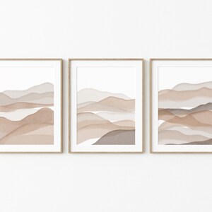 May include: Set of three framed abstract art prints featuring watercolor-style landscapes in shades of brown, beige, and gray. The prints are in simple wooden frames.