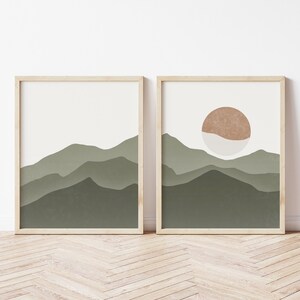 Op de afbeelding: Twee ingelijste prints met abstracte berg- en zonontwerpen. De linker print toont een groene bergketen met een witte achtergrond. De rechter print toont een groene bergketen met een witte achtergrond en een bruine zon met een wit centrum.