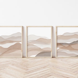 May include: Three framed prints with abstract watercolor-style landscapes in shades of brown and beige. The prints are arranged in a row on a light wood floor.