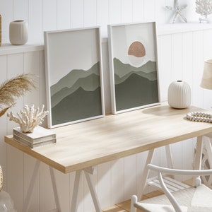 Op de afbeelding: Een wit houten bureau met twee ingelijste prints van groene en bruine bergen. Het bureau heeft een witte stoel en een witte vaas met een witte kralenketting.