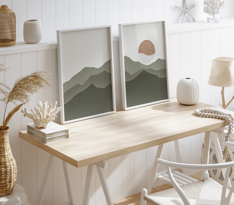 Op de afbeelding: Een wit houten bureau met twee ingelijste prints van groene en bruine bergen. Het bureau heeft een witte stoel en een witte vaas met een witte kralenketting.