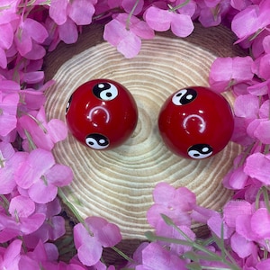 Può includere: Due oggetti rotondi rossi e lucidi, ciascuno con un simbolo yin-yang bianco e nero. Gli oggetti sono posti su uno sfondo di legno e circondati da decorazioni floreali rosa.