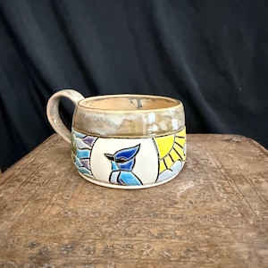 Puede incluir: Una taza de cerámica con asa ancha y borde beige. El cuerpo presenta paneles pintados a mano que representan un arrendajo azul, un sol amarillo y nubes azules sobre un fondo blanco. La taza tiene un acabado brillante y una forma redondeada.