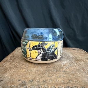 May include: A ceramic mug featuring a hand-painted black dragon on a yellow background with a blue glazed rim. The design includes a green leaf pattern on the side and a light-colored base. The mug has a rounded shape and a glossy finish.