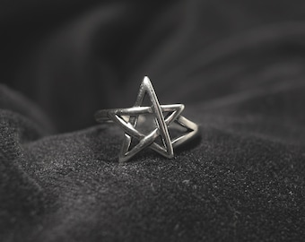 May include: A silver ring with a pentagram design. The ring is on a black fabric background.