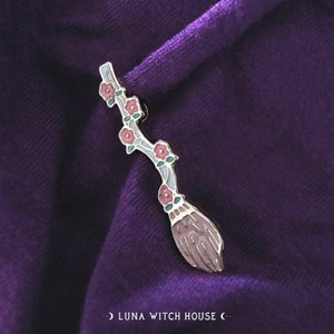May include: Gold-toned enamel pin depicting a pink and white broom with pink flowers and green leaves. The pin is displayed on a purple fabric background.  "LUNA WITCH HOUSE" is written in white text on the fabric.