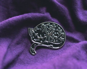 May include: A black metal pin of a sleeping cat with a crescent moon on its forehead. The cat is covered in stars and other celestial designs. The pin is on a purple fabric background.