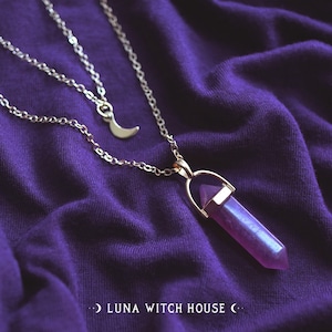 Può includere: Una collana a catena d'argento con un ciondolo a forma di luna crescente e un ciondolo di cristallo viola. La collana è appoggiata su uno sfondo di tessuto viola.