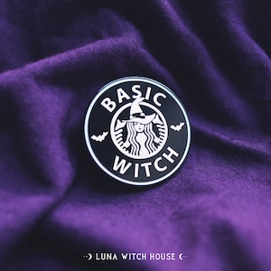 Könnte beinhalten: Runder schwarz-weißer Anstecker mit der Aufschrift "BASIC WITCH" in einem kreisförmigen Design. Der Anstecker zeigt eine Hexe mit Hut und Fledermäusen, die einem beliebten Kaffeehaus-Logo ähnelt. Auf lila Stoff.