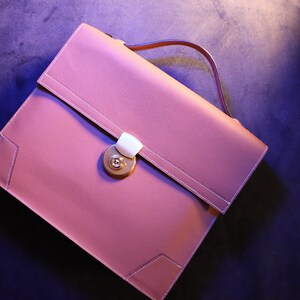May include: A dusty rose leather briefcase with a top handle and a gold-colored clasp. The briefcase has white stitching around the edges and a textured surface. The handle is brown.