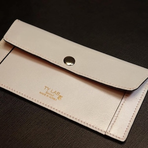 Könnte beinhalten: Eine hellrosa Ledergeldbörse mit Klappe und silbernem Druckknopf. Die Geldbörse hat schwarze Nähte und den goldenen Text "TY LAB Seoul MADE IN KOREA". Sie liegt auf einer dunklen Oberfläche.