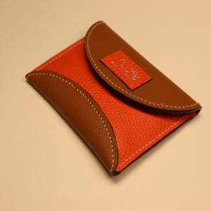 May include: A bi-color leather wallet in orange and brown with white stitching. The wallet has a curved design and a small rectangular accent on the flap. The leather has a textured appearance.