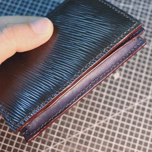 May include: A dark blue leather wallet with a textured, ridged surface. The interior is a lighter blue with red stitching. The wallet is held in a hand, showcasing the craftsmanship and design. The PDF Adobe logo is in the bottom right corner.