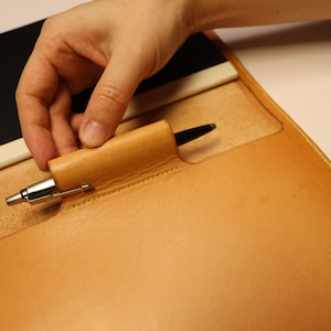 May include: A tan leather pen holder with a silver pen. The pen holder is integrated into a larger leather notebook cover. A black notebook and a white notepad are also visible.