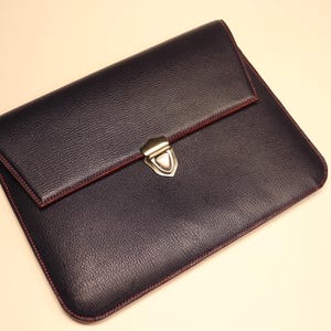 May include: A navy blue leather clutch with a textured finish. The edges are stitched with red thread, and the flap is secured with a silver-colored clasp. The interior is red.