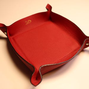 Può includere: Un vassoio rosso in pelle con forma quadrata e angoli rialzati. Il vassoio ha una superficie strutturata e un bordo nero. Gli angoli sono ripiegati e fissati con piccoli rivetti. Il vassoio ha il testo dorato "T.LAS" e "Made in Italy".