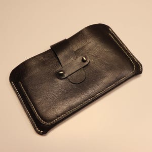 May include: A dark brown leather case with white stitching and a flap closure secured by two metal rivets. The case has a textured surface and a curved shape, possibly for a phone or small device.